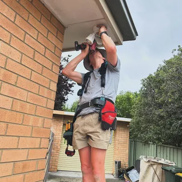 Electrician fitting a ceiling-mounted fitting under the eave of a brick Canberra home